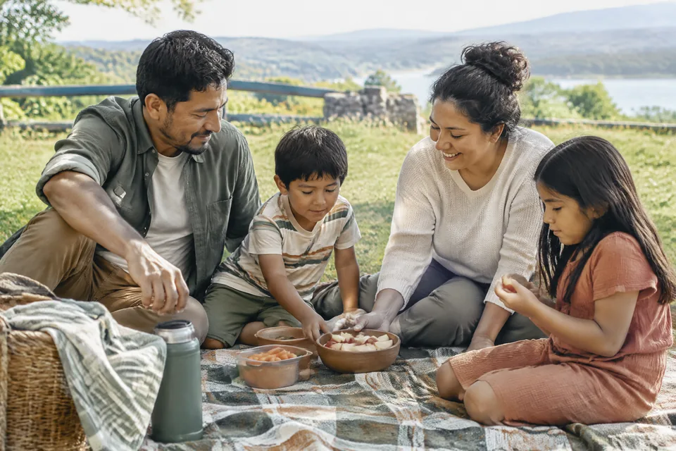 Family stretching and sharing a snack on a picnic blanket at a quiet grassy road-trip rest stop well away from cars.