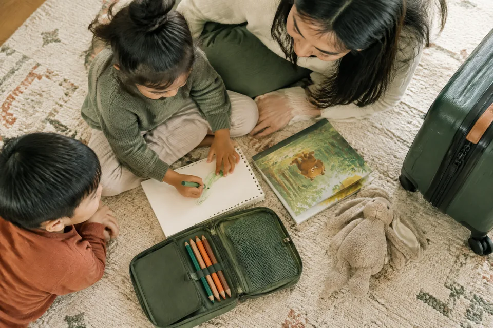 Overhead flat lay of a compact airplane activity pouch, blank sketchpad, thick pencils, picture-only paperback and one soft toy beside a carry-on.